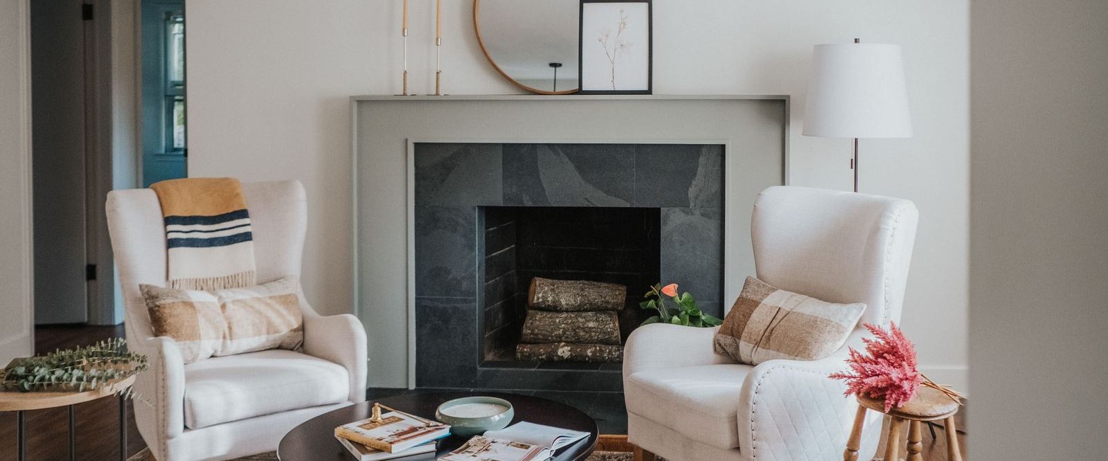 Living room with two cream wingback chairs facing a slate fireplace, a round wood mirror on the mantel